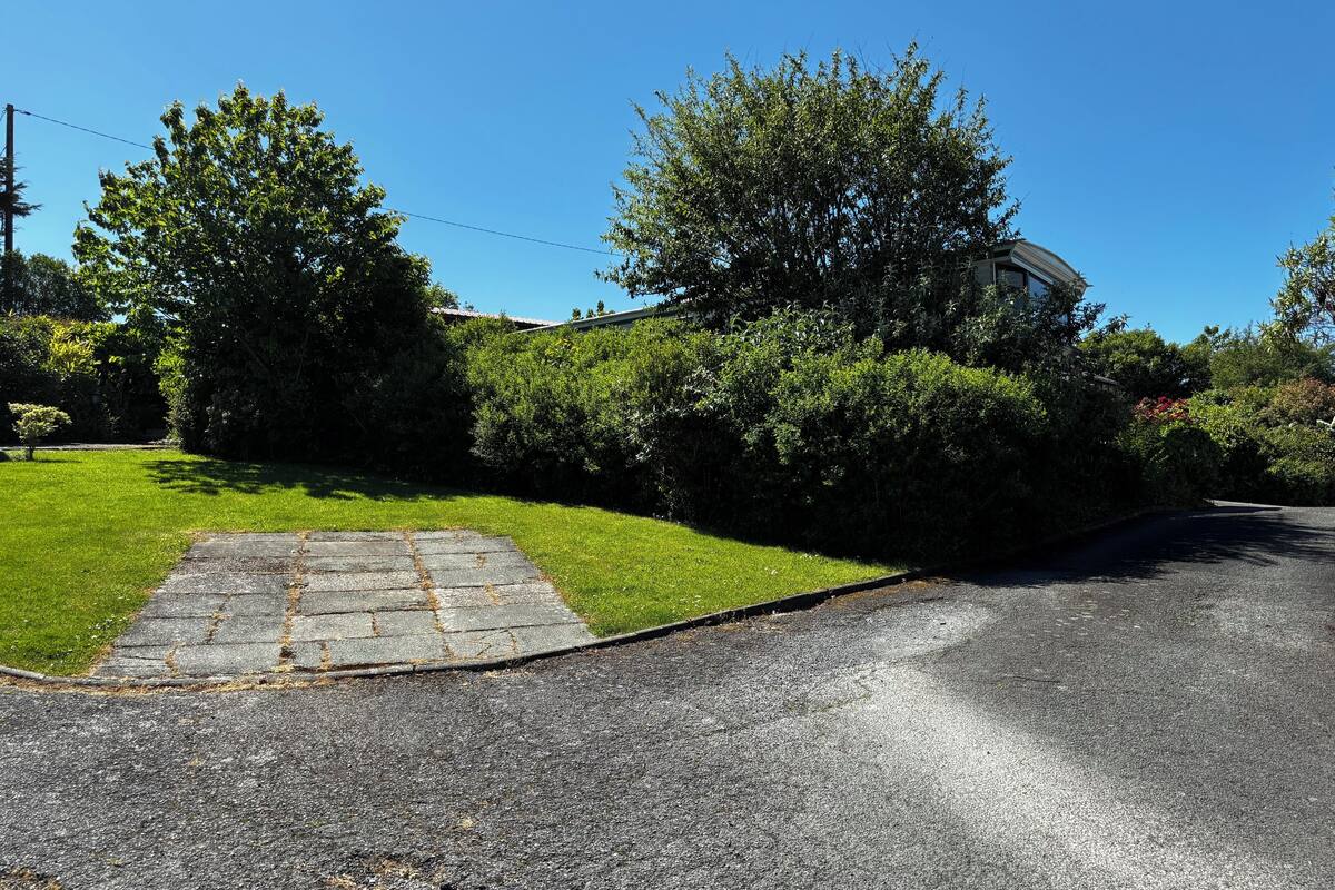 A paved driveway leading to a house with a sloped roof, surrounded by lush greenery and a clear blue sky.