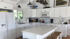 Une cuisine moderne avec des meubles blancs, un îlot central doté de tabourets, une poubelle en acier inoxydable et une fenêtre laissant entrer la lumière naturelle.