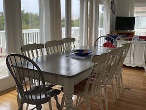 Un coin repas avec une table en bois et des chaises blanches, un saladier posé sur la table, et un téléviseur sur un meuble en arrière-plan.