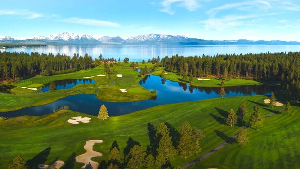 Un campo de golf con un lago, bosque y montañas al fondo.