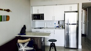 A compact kitchen with white cabinets, a stainless steel refrigerator, a microwave, and a sink. There is a small stool and a dark-colored sofa with a decorative pillow. - Newly remodeled beach front! SPECIAL RATE SEPTEMBER and OCTOBER (Marco Island)