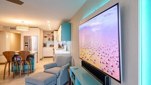 Ein modernes Wohnzimmer mit einem großen Flachbildfernseher an der Wand, einem gestreiften Sessel, einem kleinen Couchtisch und einem Essbereich mit Küche im Hintergrund.