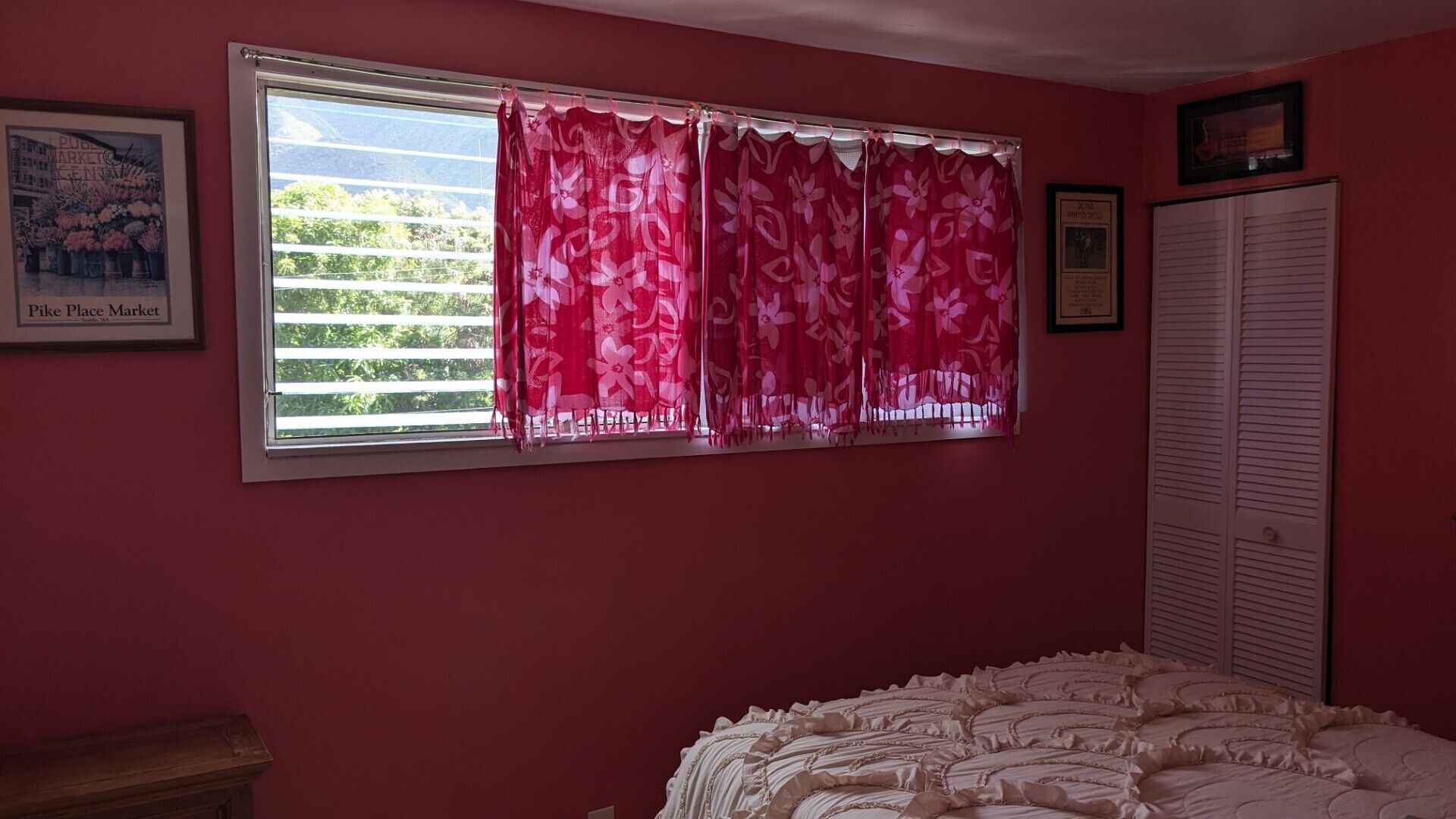 A bedroom with a large window, a bed with a white bedspread, a wooden nightstand, and a framed picture on the wall.