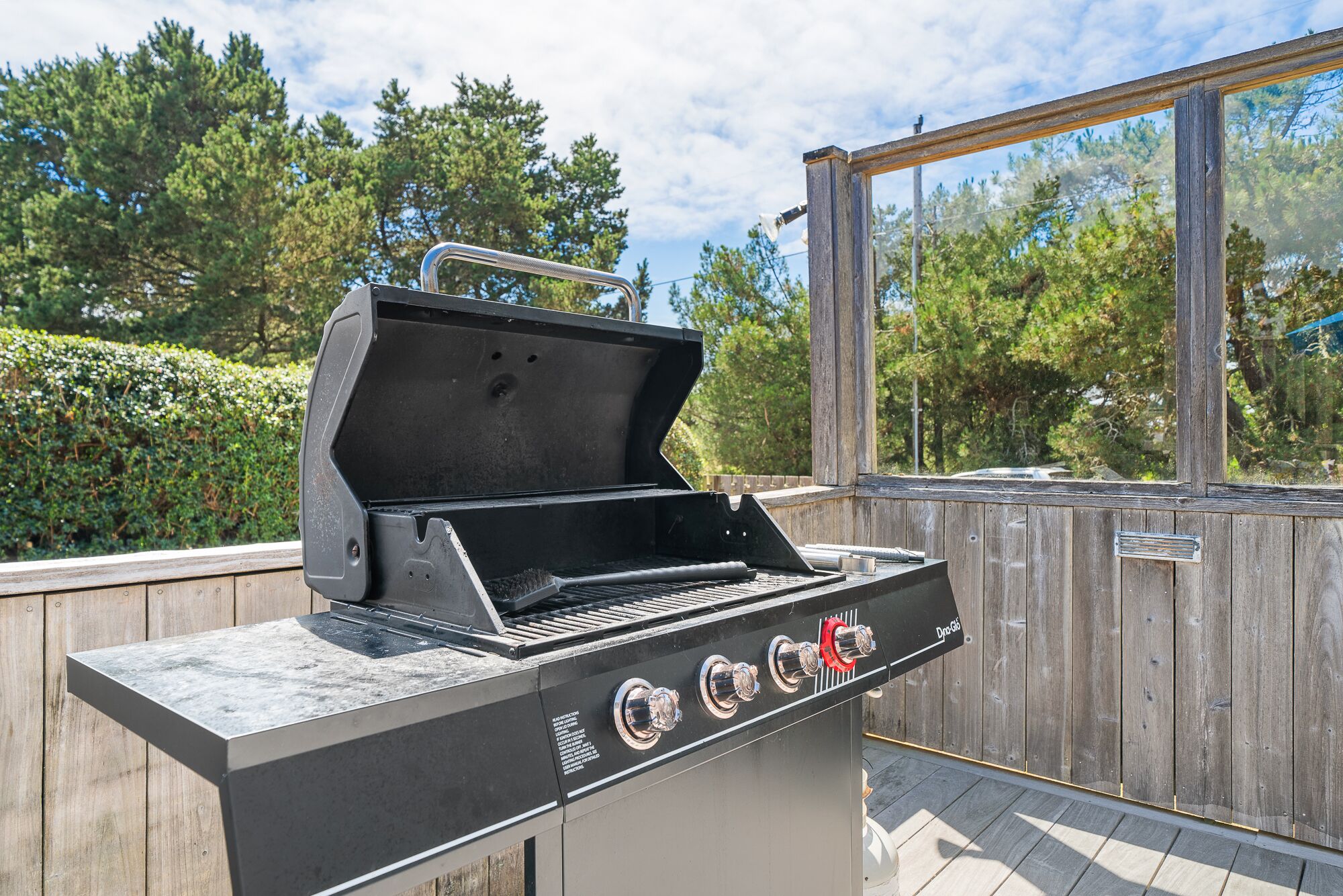 A barbecue grill with four burners, a side shelf, and a window overlooking a wooded area.