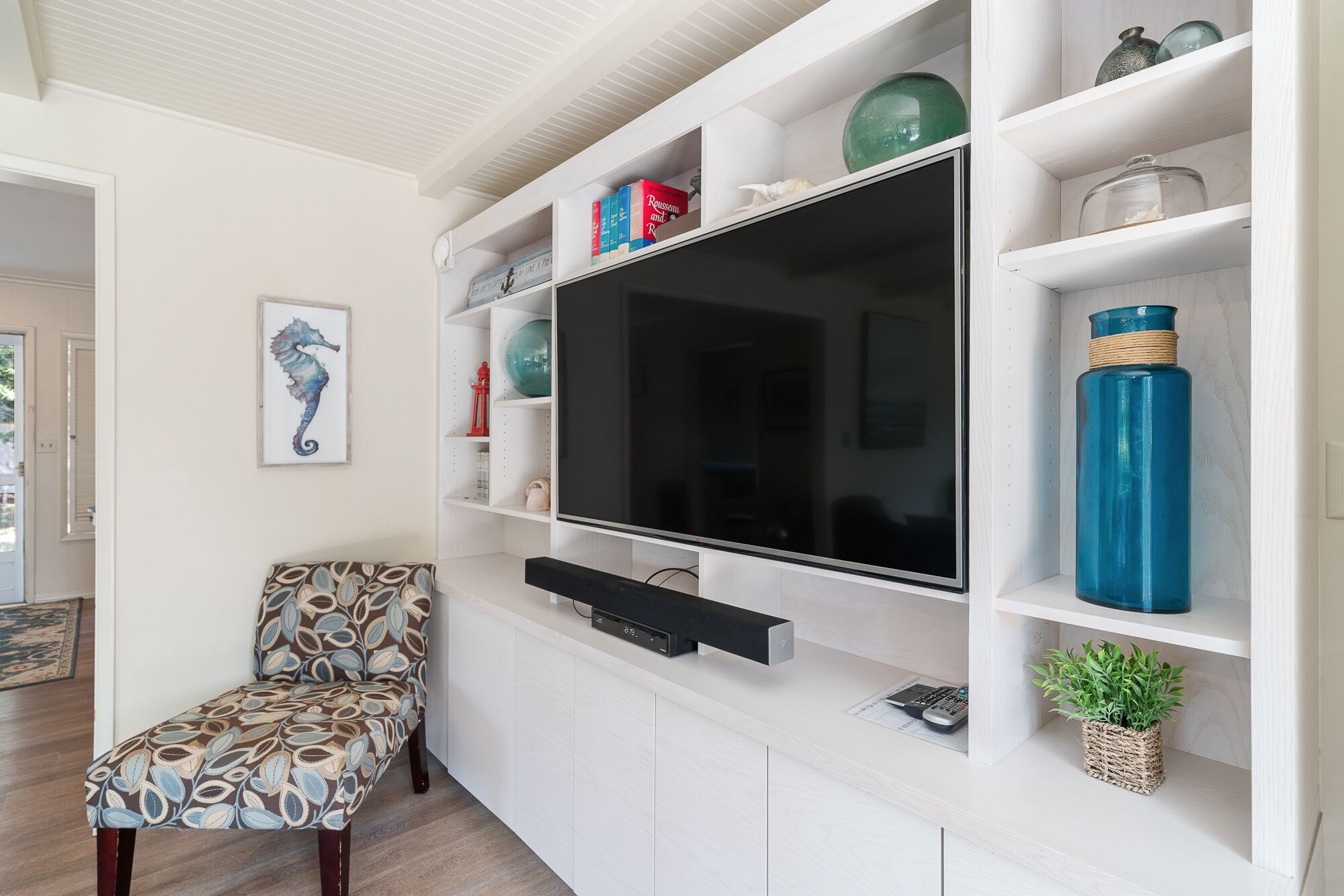 Ein Wohnzimmer mit Fernseher an einer weißen Wand, einem gemusterten Sessel und Regalen mit Dekoration.
