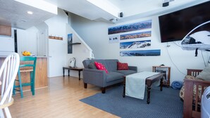 Ein Wohnzimmer mit grauem Sofa, einem Holztisch, einem blauen Teppich und einem flachbildfernseher, der an der Wand befestigt ist.