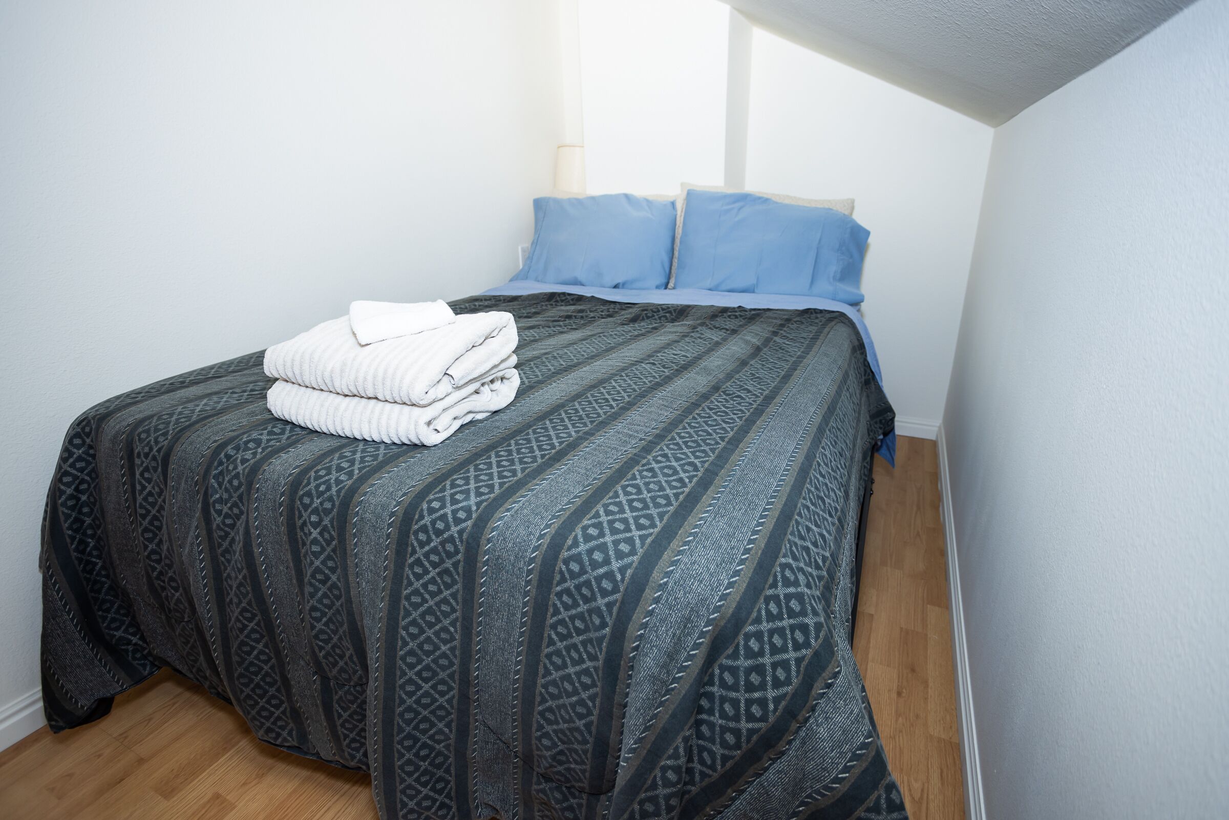 A neatly made bed with a patterned bedspread and two pillows, a folded white towel, and a wooden floor.
