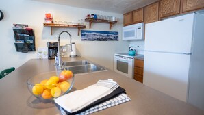 Eine Küche mit Spüle aus Edelstahl, Wasserkocher, Mikrowelle und Kühlschrank. Darüber befinden sich Holzschränke und ein Regal mit Gläsern und einer Kaffeemaschine.