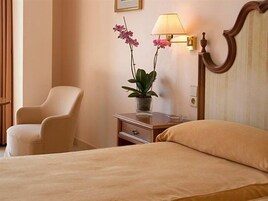 Una habitación de hotel con una cama, una silla, una mesita de noche con lámpara y una planta en maceta.