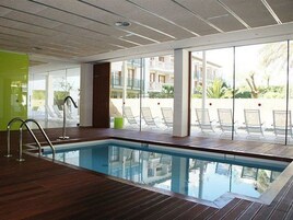 Piscina interior con paredes de vidrio transparente, terraza de madera y vistas al exterior.
