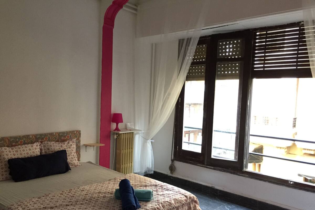 A bedroom with a bed, a nightstand, a window with blinds, and a view of the outside.