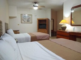 Habitación de hotel con dos camas, un armario de madera, un escritorio con lámpara, un ventilador de techo y un cuadro en la pared.