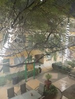 Un área de estar al aire libre con mesas y sillas, rodeada de follaje y un edificio con un letrero verde.