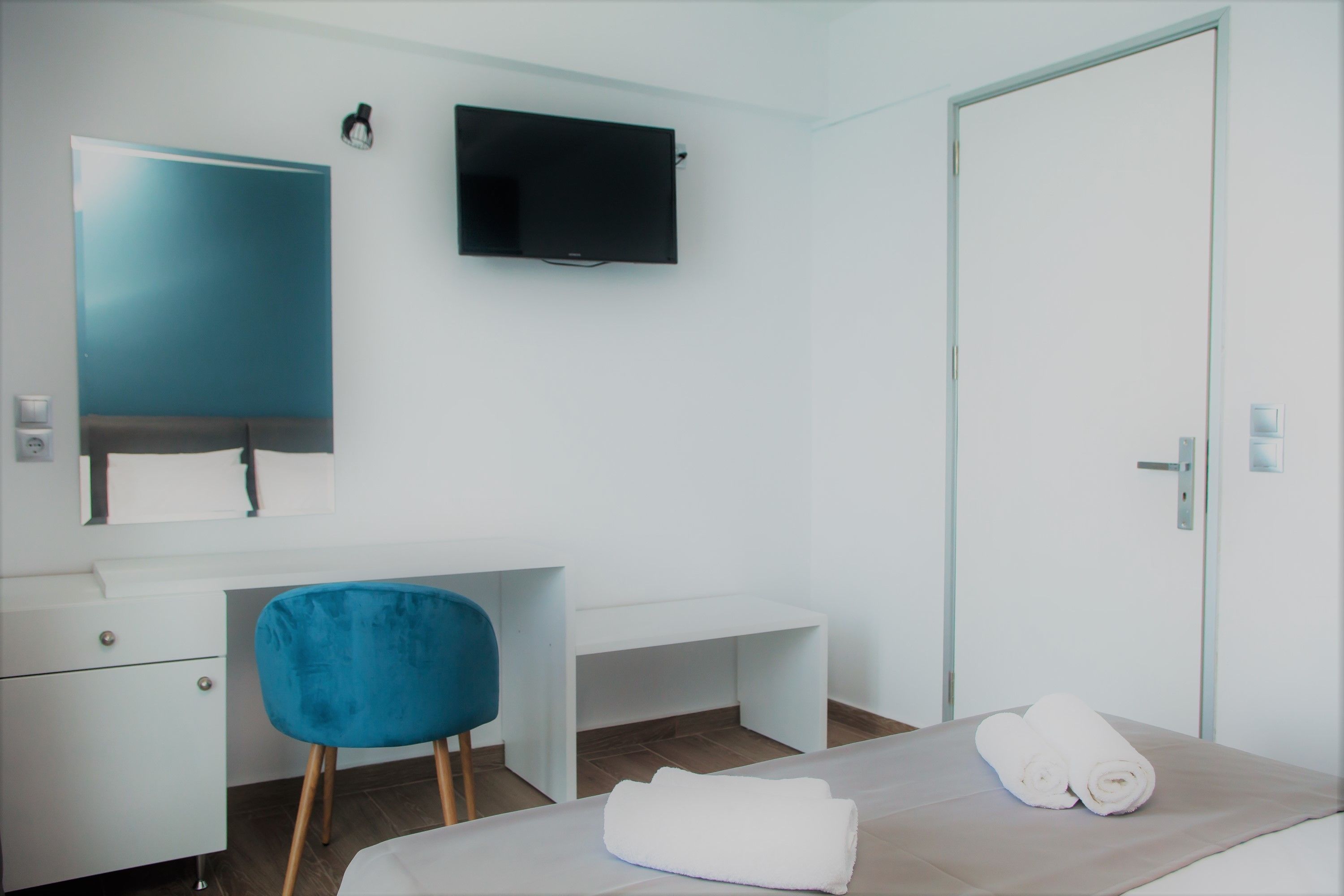 Una camera d'albergo con un letto, una poltrona blu, una scrivania con uno specchio, una televisione e una porta.