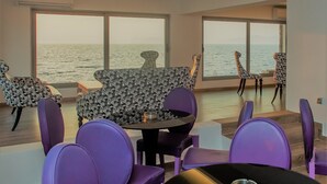 A dining area with purple chairs and a table, large windows with a view of the sea, and patterned chairs. - Sunrise Aparthotel Rhodes (Rhodes)