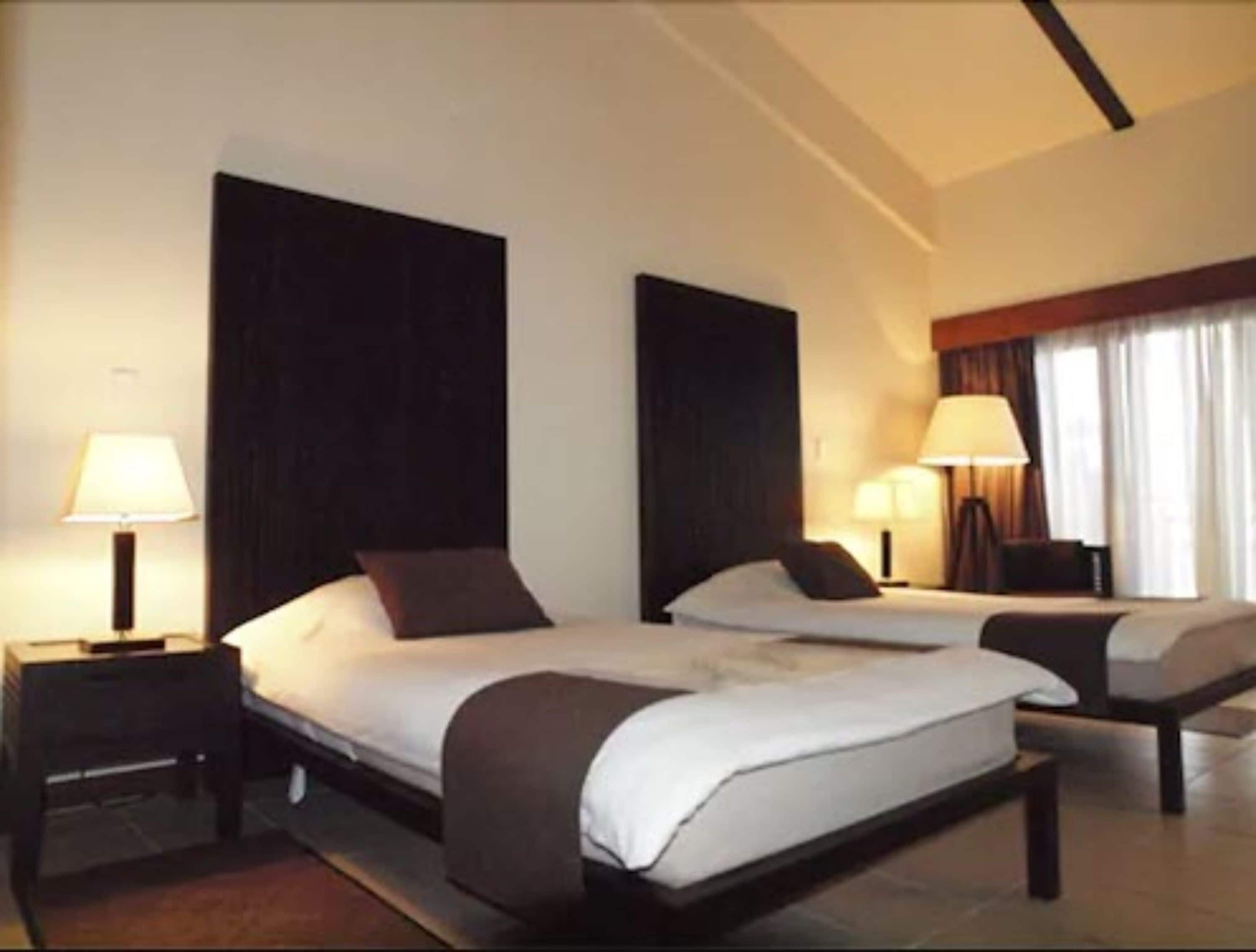 A hotel room with two beds, dark wood headboards, bedside lamps, and a large window with curtains.
