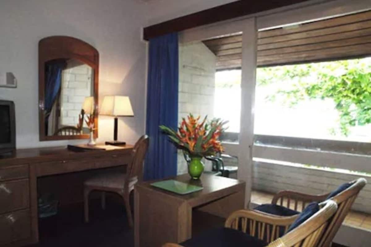 A hotel room with a wooden desk, two chairs, a lamp, a mirror, and a window with blue curtains.