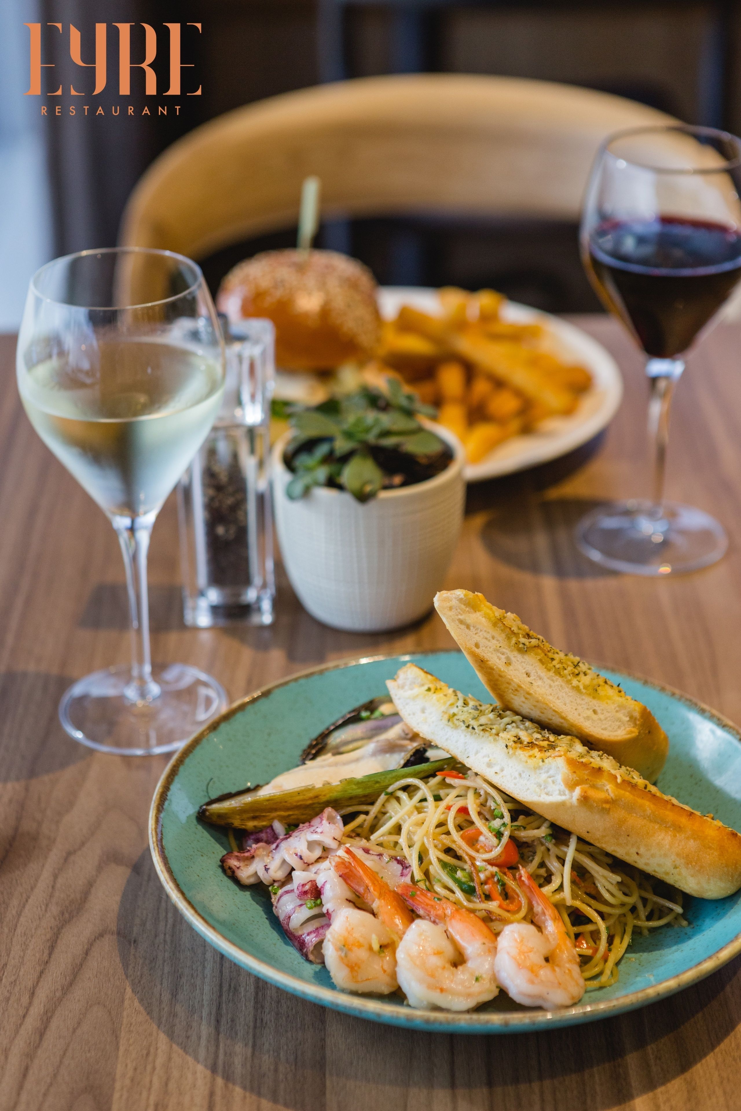 A dining table with a plate of pasta, shrimp, and bread, a glass of white wine, and a bowl of fries.