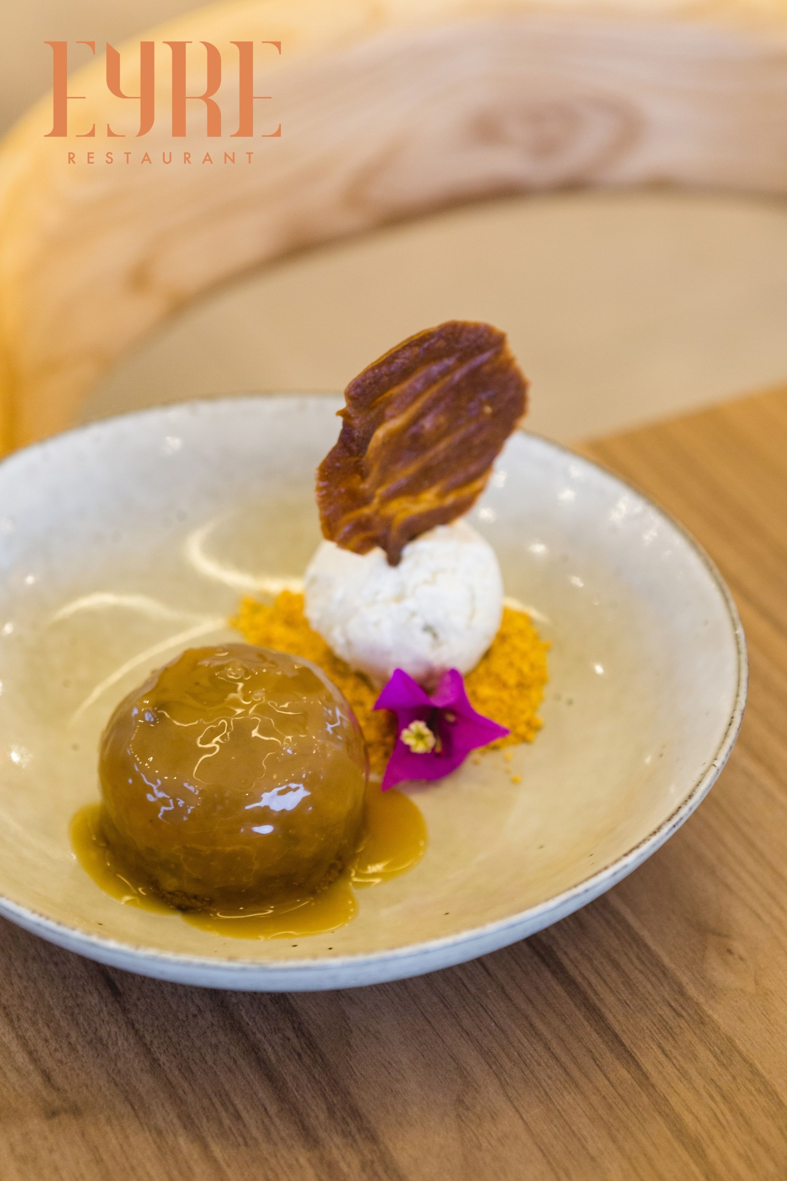 A dessert plate with a caramel sphere, a white scoop, and a decorative leaf on a ceramic plate.