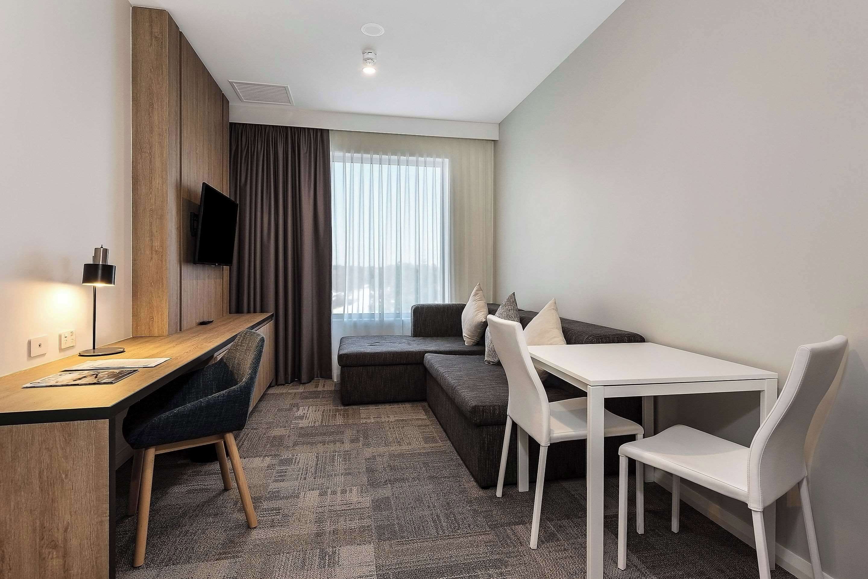Una habitación de hotel moderna con un escritorio de madera, un televisor de pantalla plana, un sofá y una mesa de comedor con sillas.