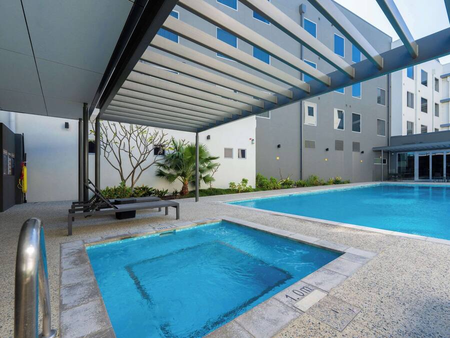 Área de piscina com espaço de estar coberto, posto de salva-vidas e um prédio ao fundo.