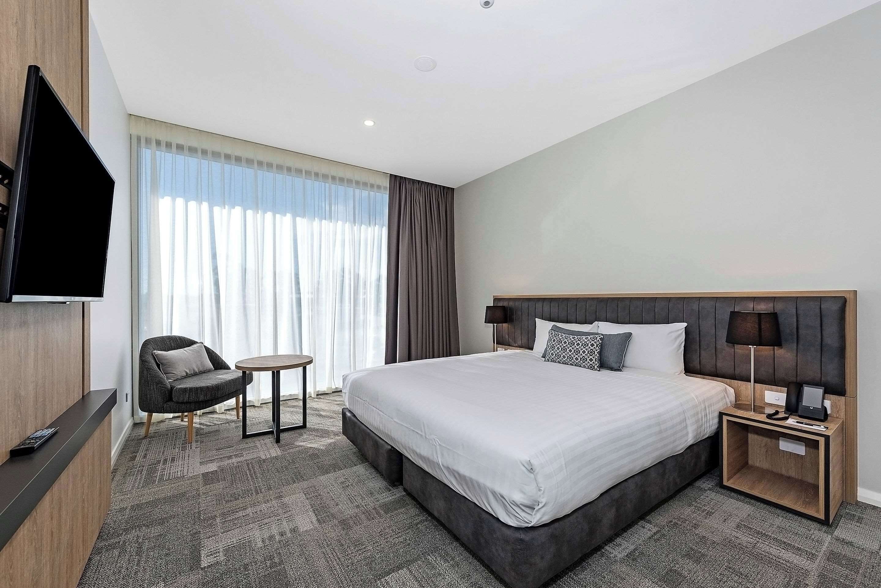Una habitación de hotel moderna con una cama grande, un televisor de pantalla plana montado en la pared, una mesita redonda y un rincón de descanso con una silla.