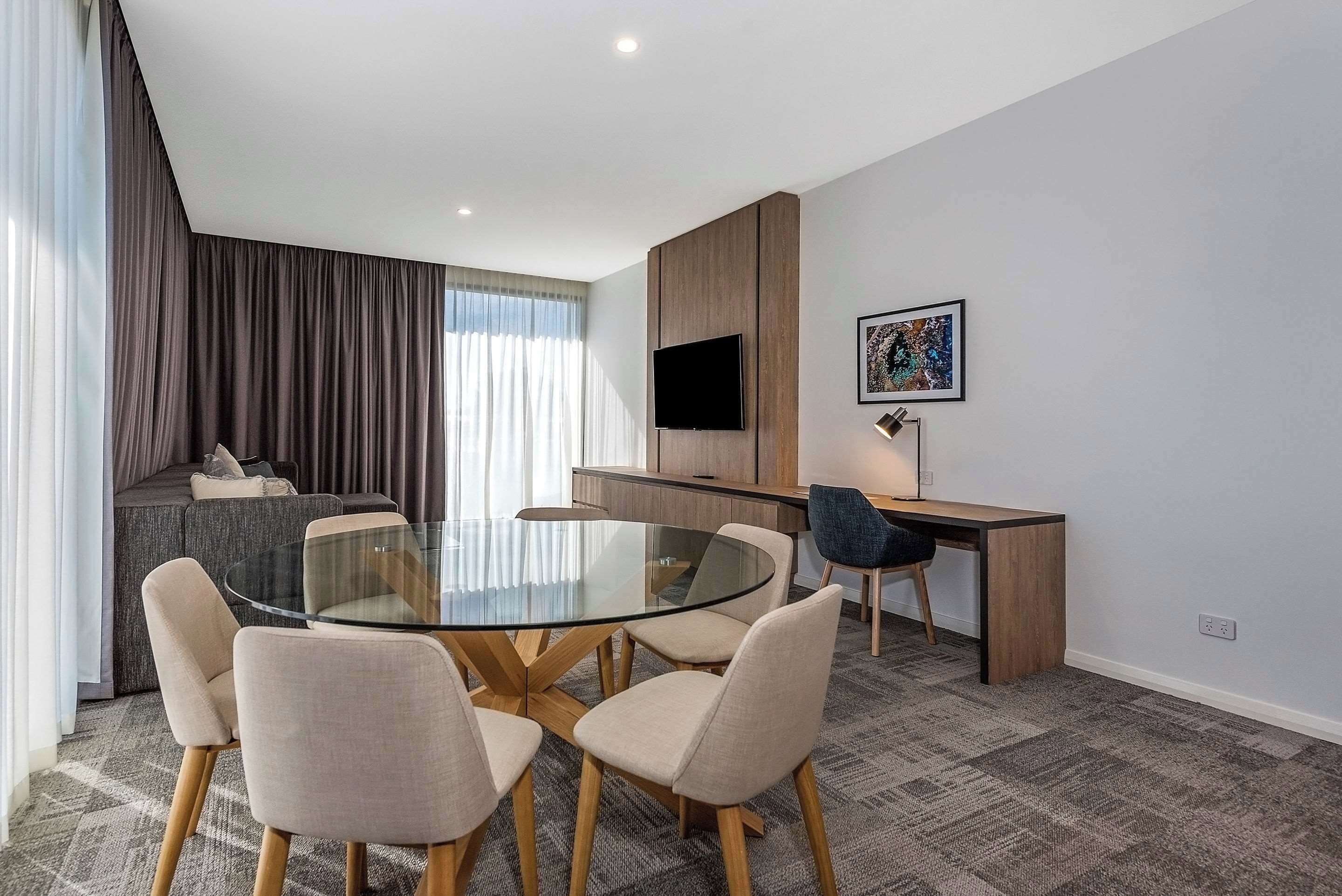 Una habitación de hotel moderna con una mesa redonda de vidrio, seis sillas, un sofá, un escritorio, un televisor de pantalla plana y una obra enmarcada en la pared.