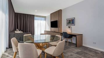 Una habitación de hotel moderna con una mesa redonda de vidrio, seis sillas, un sofá, un escritorio, un televisor de pantalla plana y una obra enmarcada en la pared.