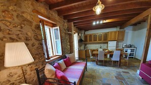 A cozy room with a stone wall, a bed with pillows, a table with a lamp, and a view of a kitchen with wooden cabinets and a dining area. - Villa Kalata (Lefkada)