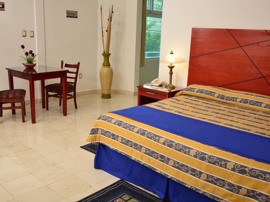 Habitación de hotel con cama, escritorio, sillas, un florero con flores, lámpara y ventana.