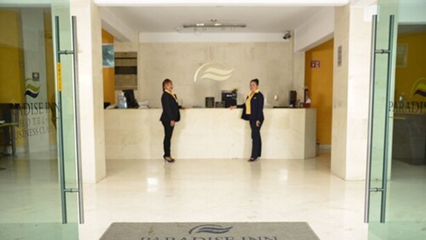 Et receptionsområde med to receptionister ved en disk, et logo over disken, og en dørmåtte med hotellets navn.