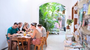 A group of people dining at a wooden table in a room with a white wall and an archway leading to a garden. - Tunido - Hostel (Santa Marta)