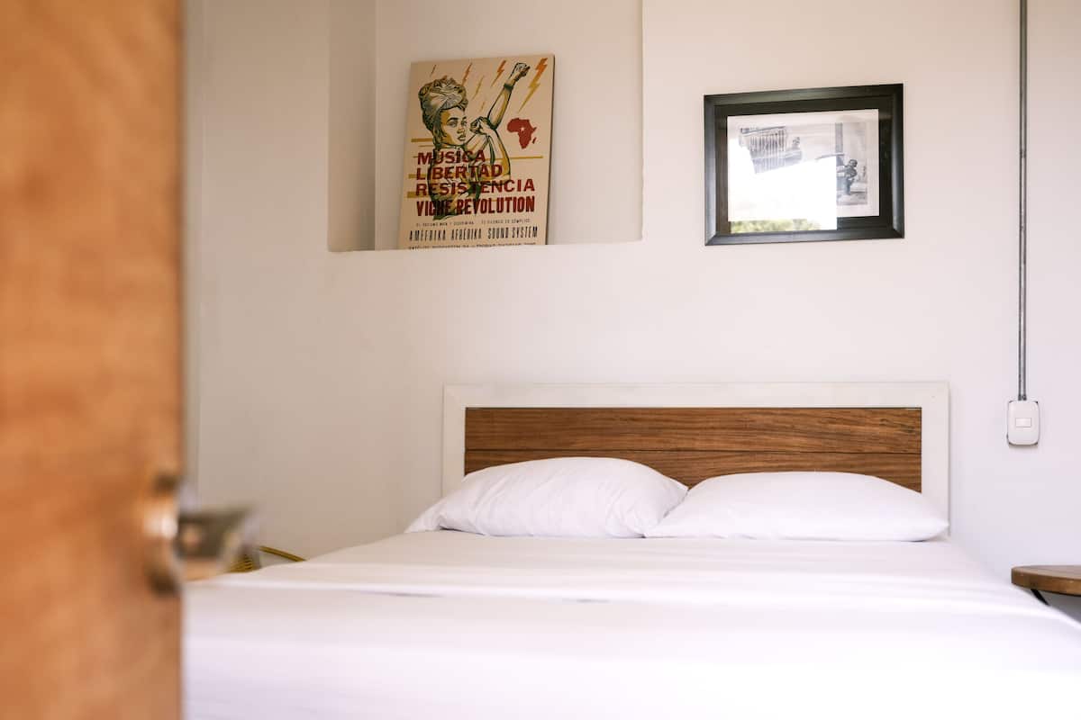 A neatly made bed with white linens in a room with a wooden headboard. There is a framed picture on the wall above the bed and a poster with text and graphics.