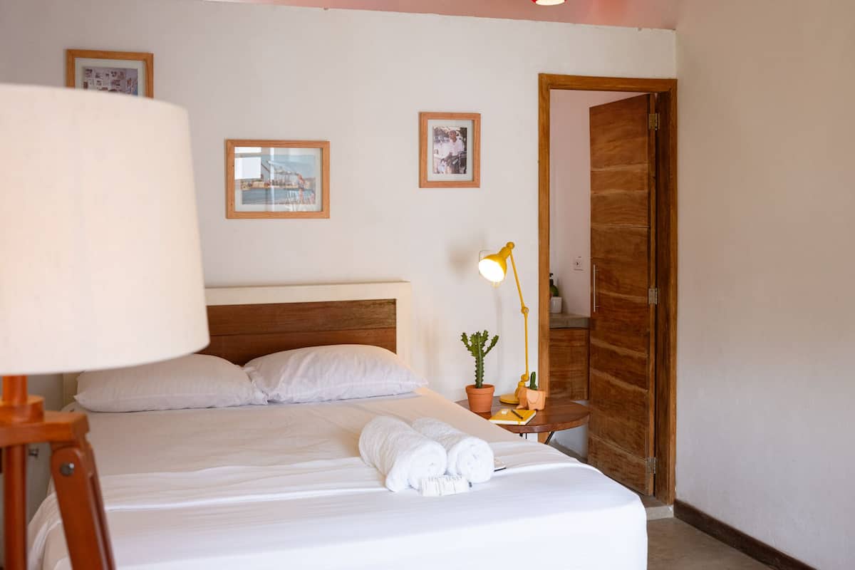 A neatly made bed with white linens and rolled towels, a wooden headboard, two framed pictures on the wall, a small table with a lamp and a potted plant, and a wooden door in the background.
