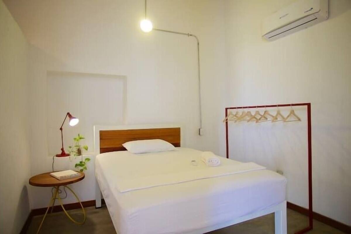 A neatly made bed in a room with a bedside table, a hanging clothes rack, and an air conditioner on the wall.