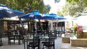An outdoor dining area with tables and chairs under blue umbrellas, surrounded by trees and a brick planter with flowers. - Hotel Les Cigalons (Marseille)