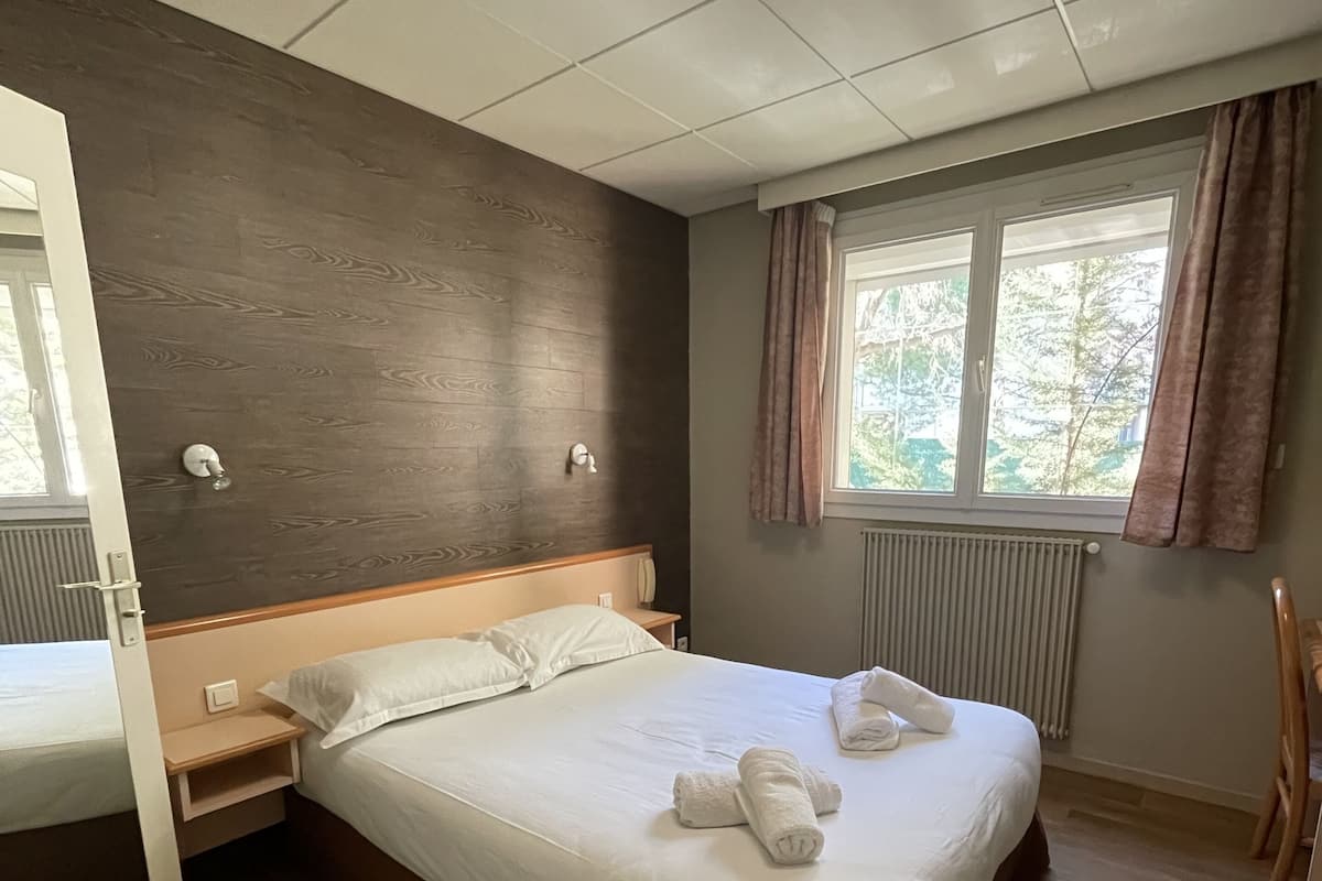 Une chambre d’hôtel avec un lit, des tables de chevet, une fenêtre avec des rideaux, un radiateur et un plancher en bois.