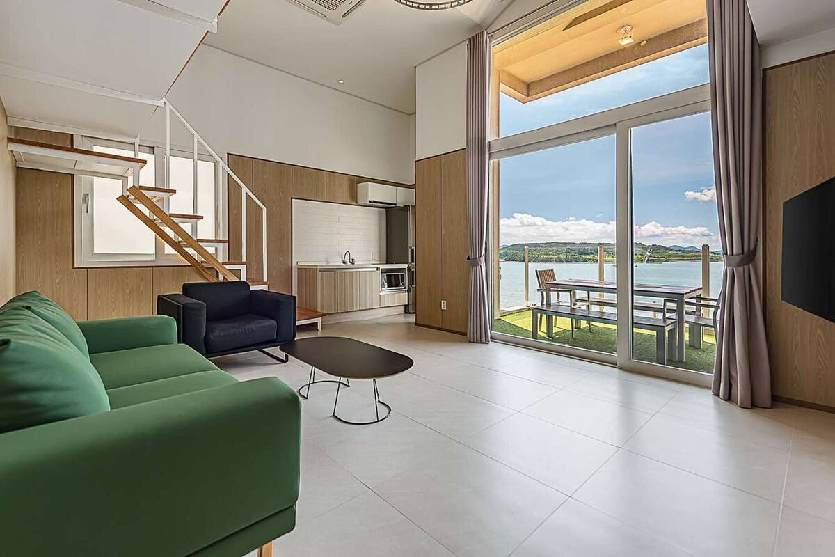 Ein modernes Wohnzimmer mit einem grünen Sofa, einem schwarzen Sessel und einem Essbereich mit Tisch und Stühlen. Eine Küche mit Holzausstattung und eine Treppe sind ebenfalls zu sehen.