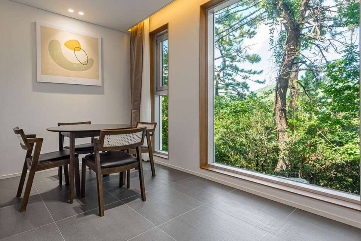 Ein Essbereich mit Tisch und Stühlen, ein großes Fenster mit Blick auf einen Wald und ein abstraktes Kunstwerk an der Wand.
