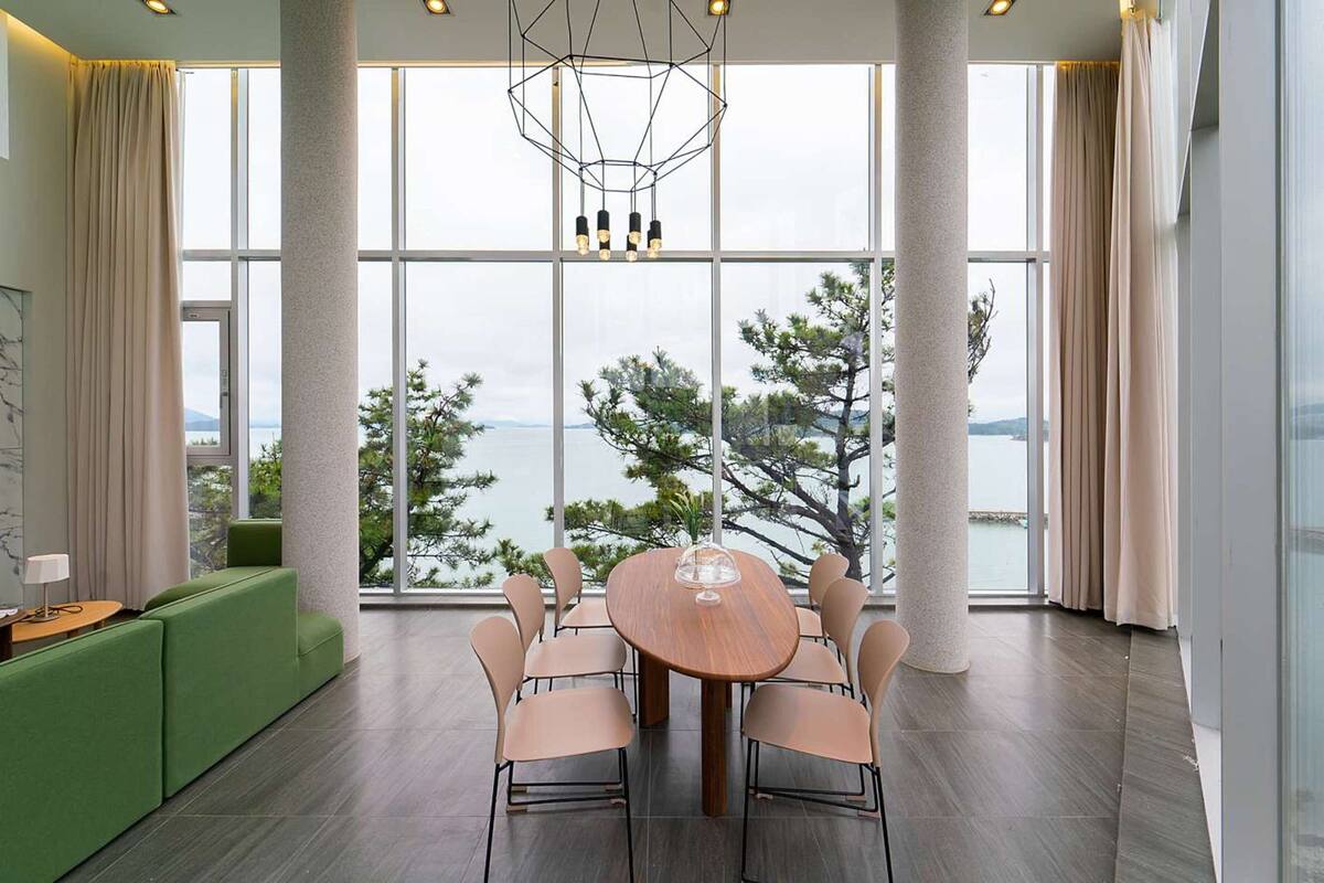 Ein moderner Essbereich mit einem runden Tisch und Stühlen, großen Fenstern mit Blick auf Bäume und Wasser sowie einem grünen Sofa.