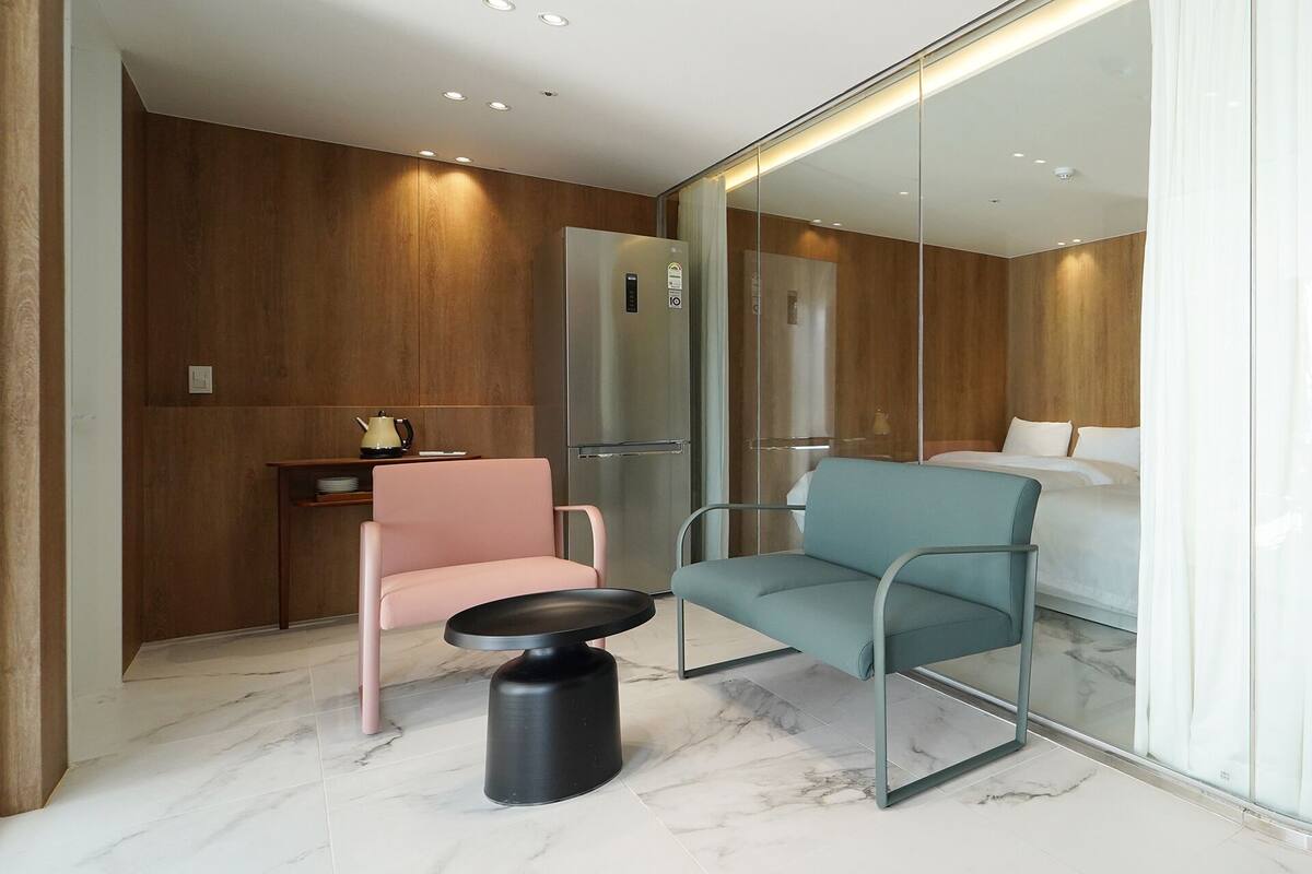 Ein modernes Hotelzimmer mit einer Holzwand, einem Bett, einem rosa Sessel, einem türkisblauen Sessel und einem kleinen Tisch mit Wasserkocher.