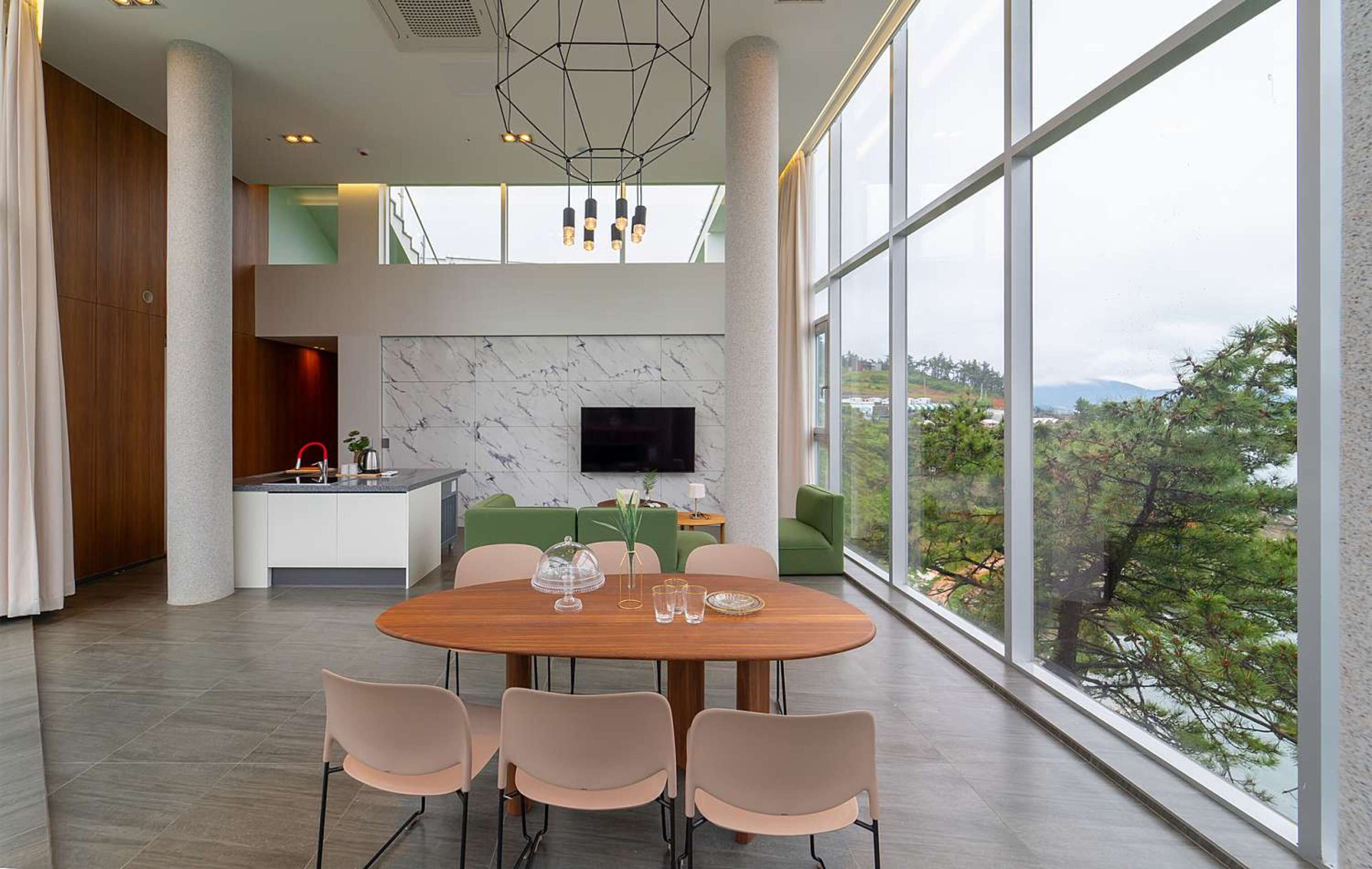 A modern dining area with a wooden table and chairs, a kitchen in the background, and a large window with a view of trees and mountains.
