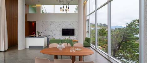 A modern dining area with a wooden table and chairs, a kitchen in the background, and a large window with a view of trees and mountains.