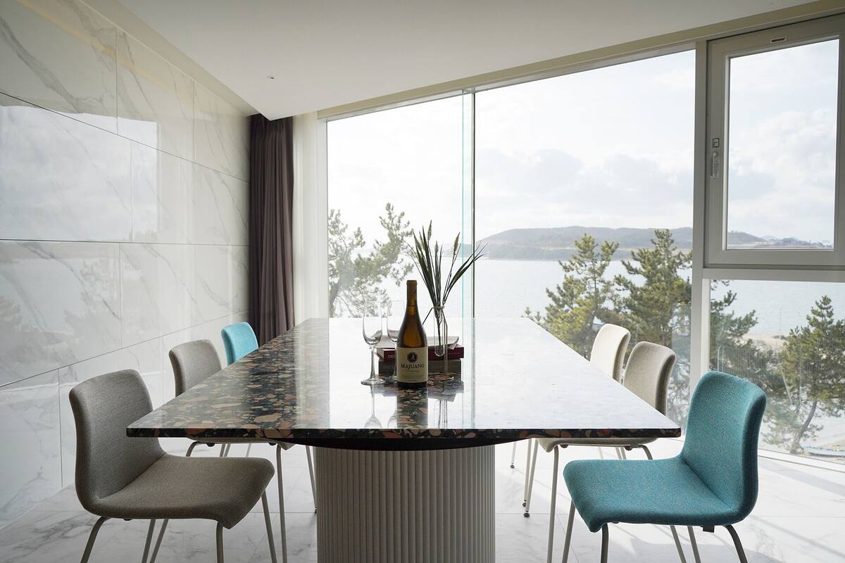 Ein moderner Essbereich mit einem Marmortisch, vier Stühlen und Blick auf einen See durch große Fenster.