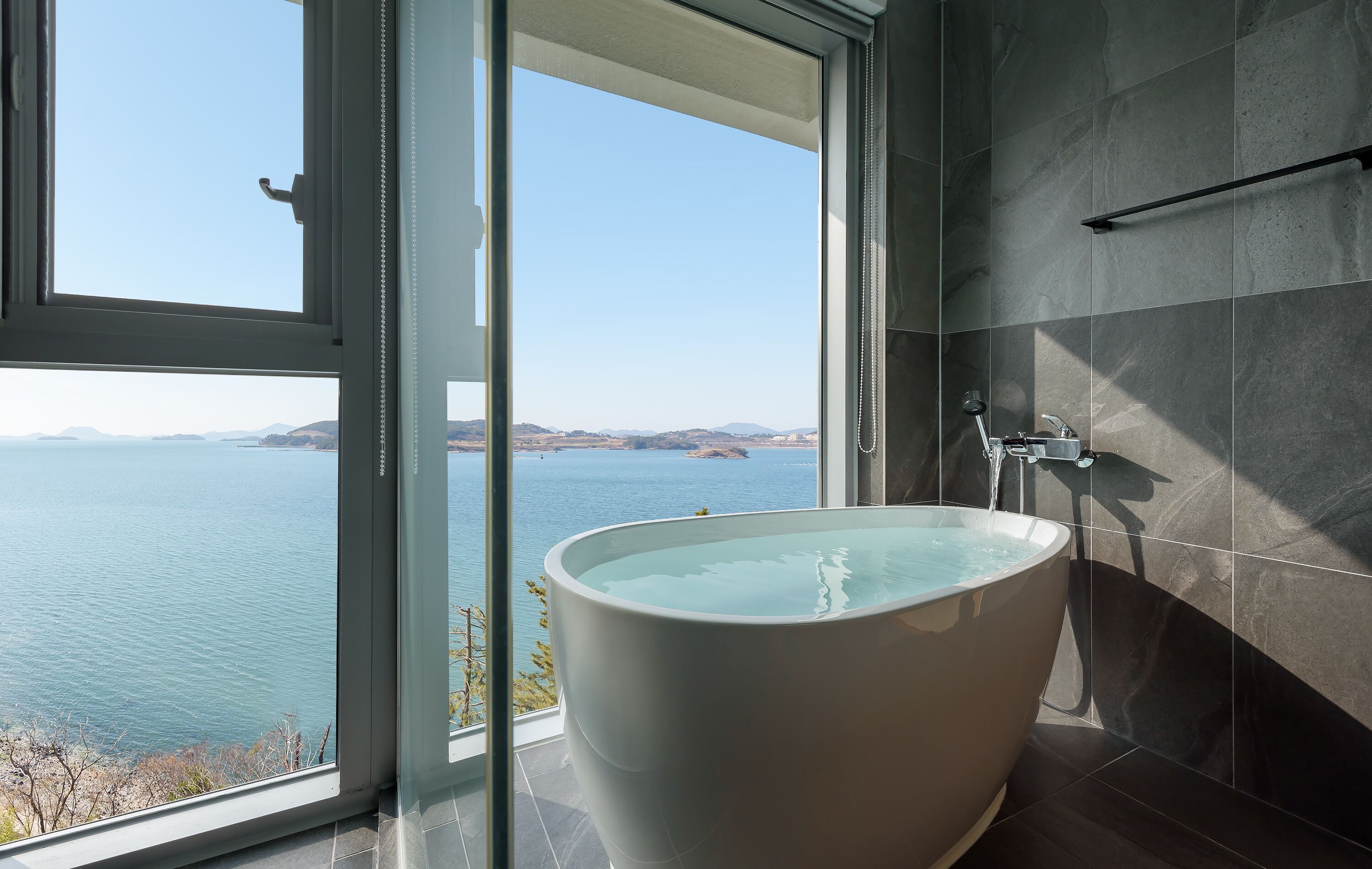 Un baño moderno con una bañera grande, ducha y vista al mar a través de un ventanal amplio.
