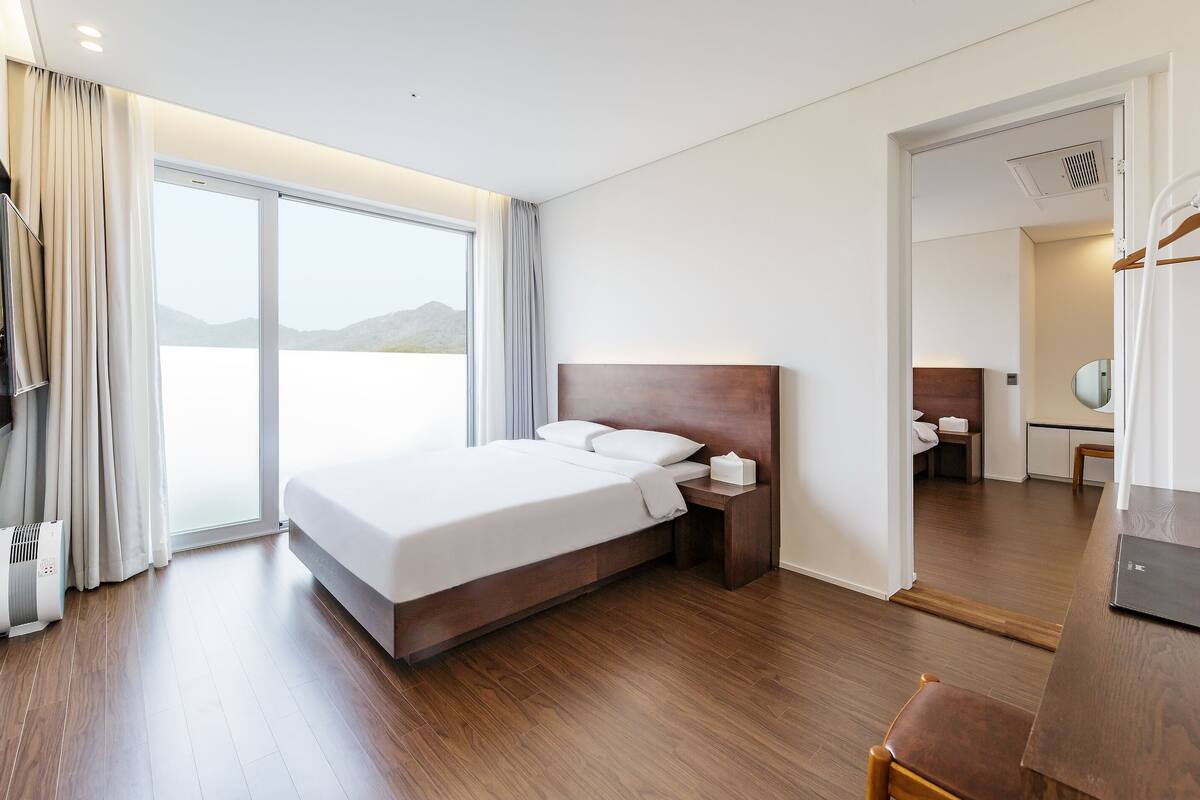 Ein modernes Hotelzimmer mit einem großen Bett, einem Nachttisch, einem Schreibtisch mit Stuhl, einem Spiegel und Blick ins Freie.
