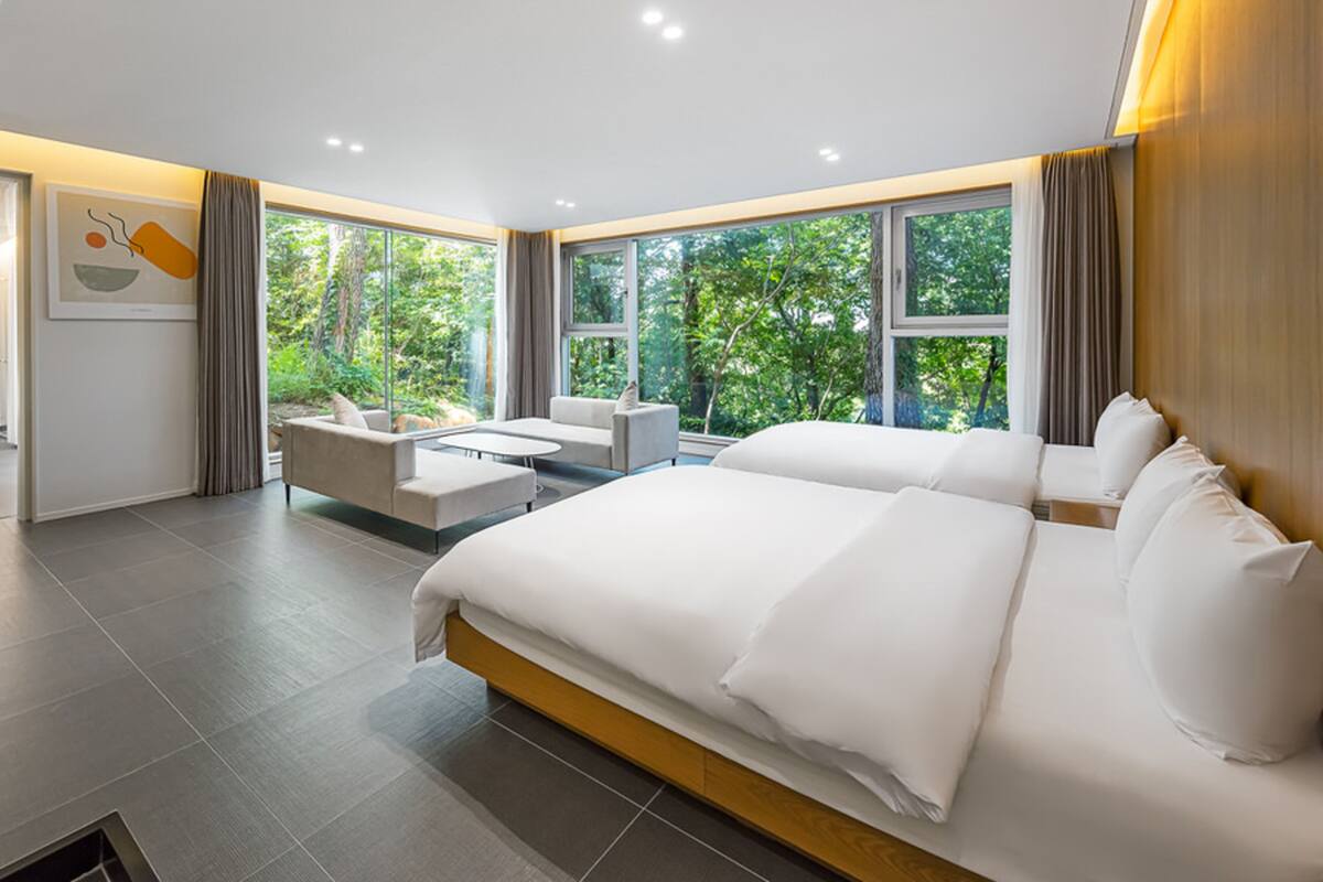 Ein modernes Schlafzimmer mit einem großen Bett, einer Couch und einem Couchtisch. Große Fenster mit Vorhängen und Blick auf Bäume.