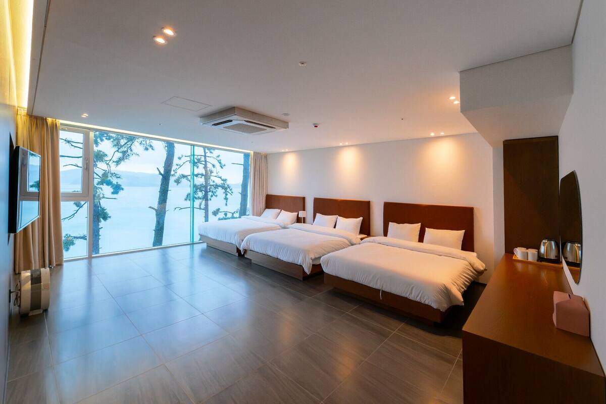 Ein modernes Hotelzimmer mit zwei Betten, einem großen Fenster mit Blick auf Bäume und Wasser sowie einem Holztisch mit Spiegel und Kaffeemaschine.