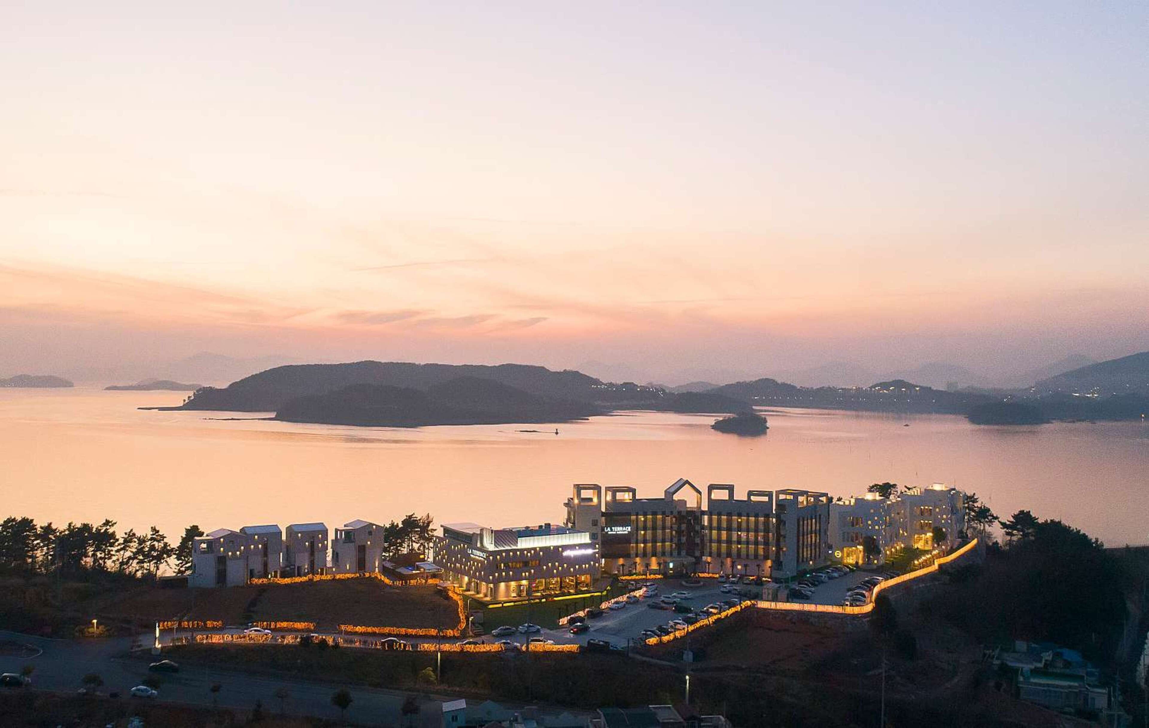 A coastal hotel with modern architecture, a marina, and a view of the sea and distant hills.