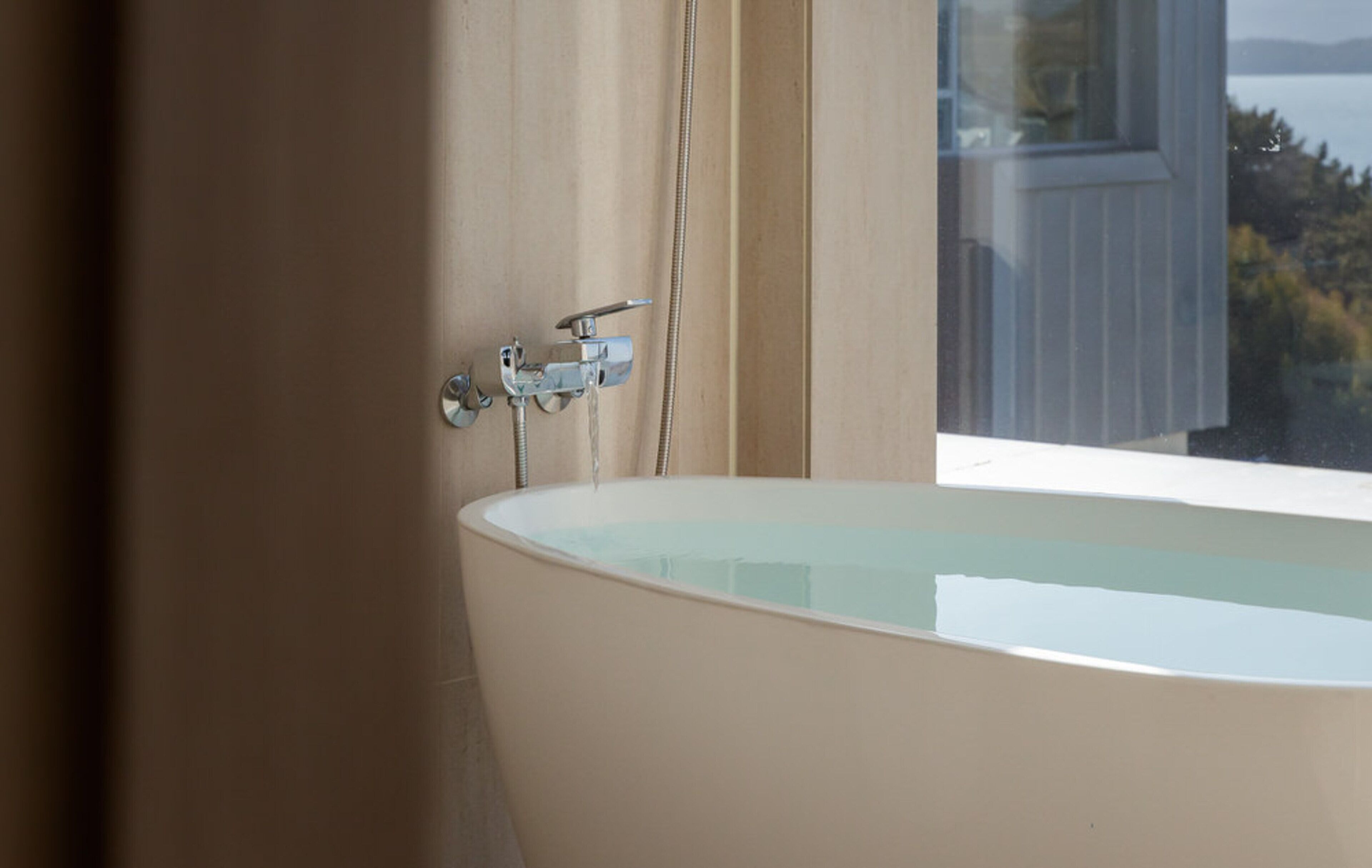A bathtub with a showerhead, a window with a view of trees and a building, and a curtain.